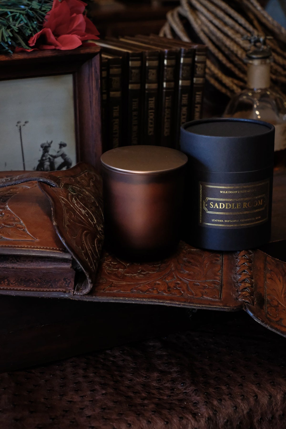 Wilkinson's Fine Goods Saddle Room candle beside its black paper tube on a hand tooled leather tray, with leather bound books and a coiled rope behind.