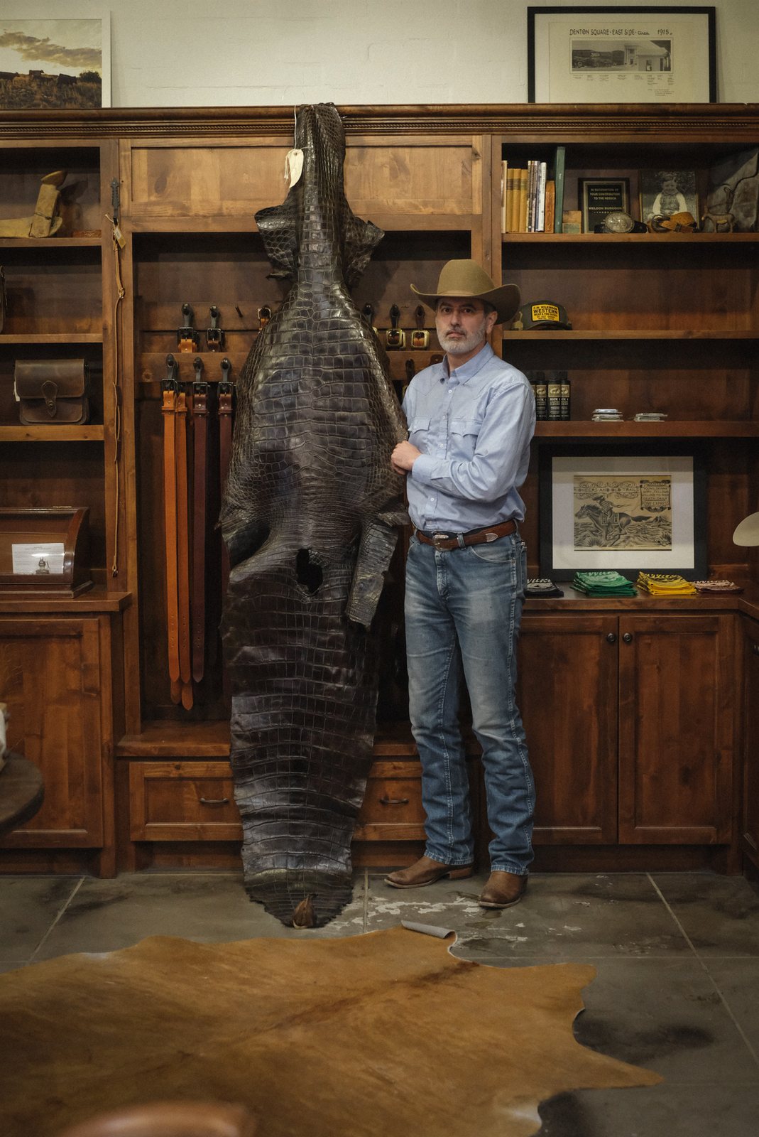 A man in a cowboy hat, light blue shirt, and jeans stands next to a large, taxidermy-alligator-style hide mounted on a wooden wall display in a rustic room with bookshelves.