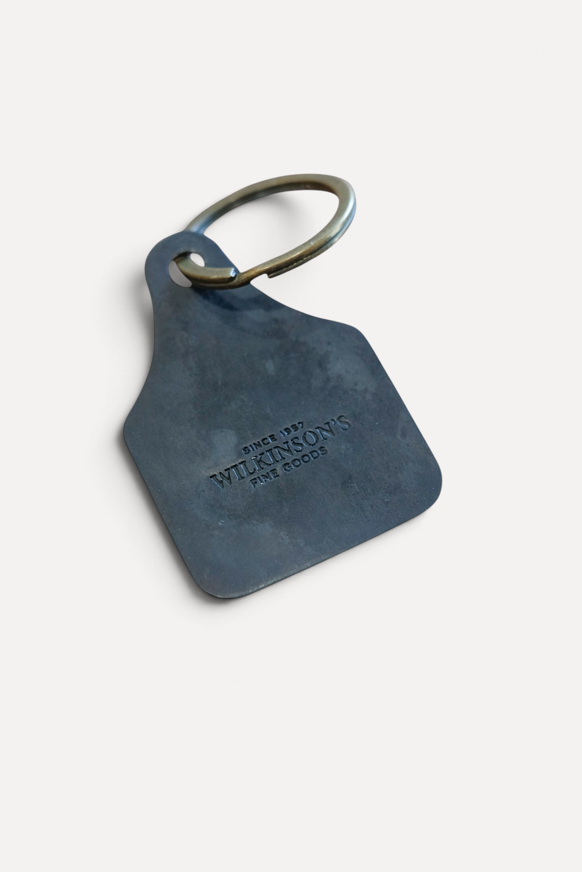 Wilkinson's Fine Goods custom steel stock tag with brass ring on a wooden surface